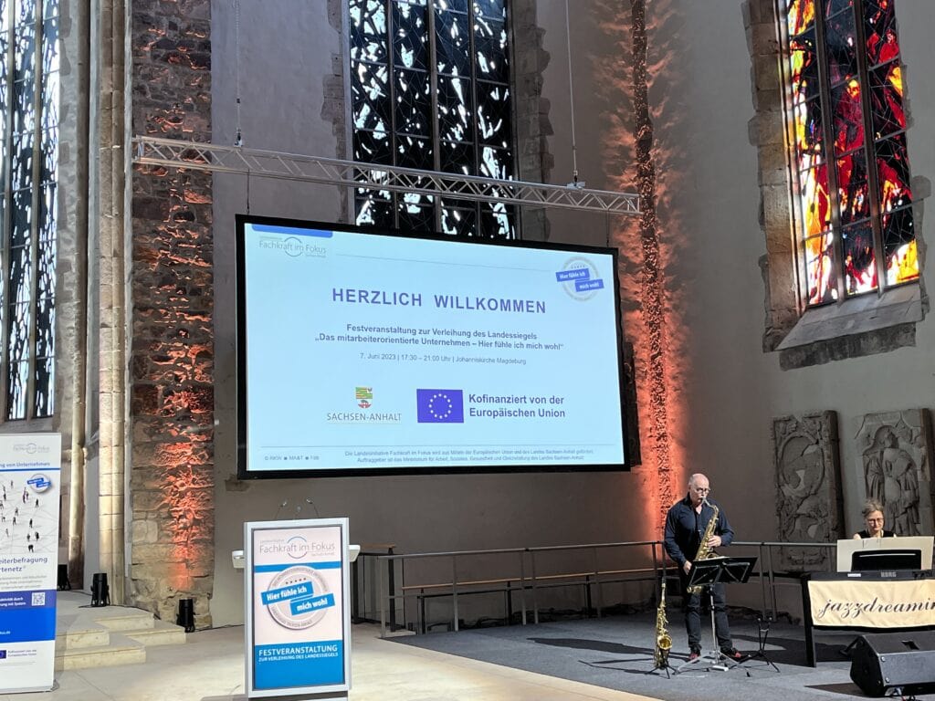 Veranstaltung zur Landessiegel-Verleihung. Großer Willkommens-Bildschirm und ein Mann spielt davor ein Blasinstrument.