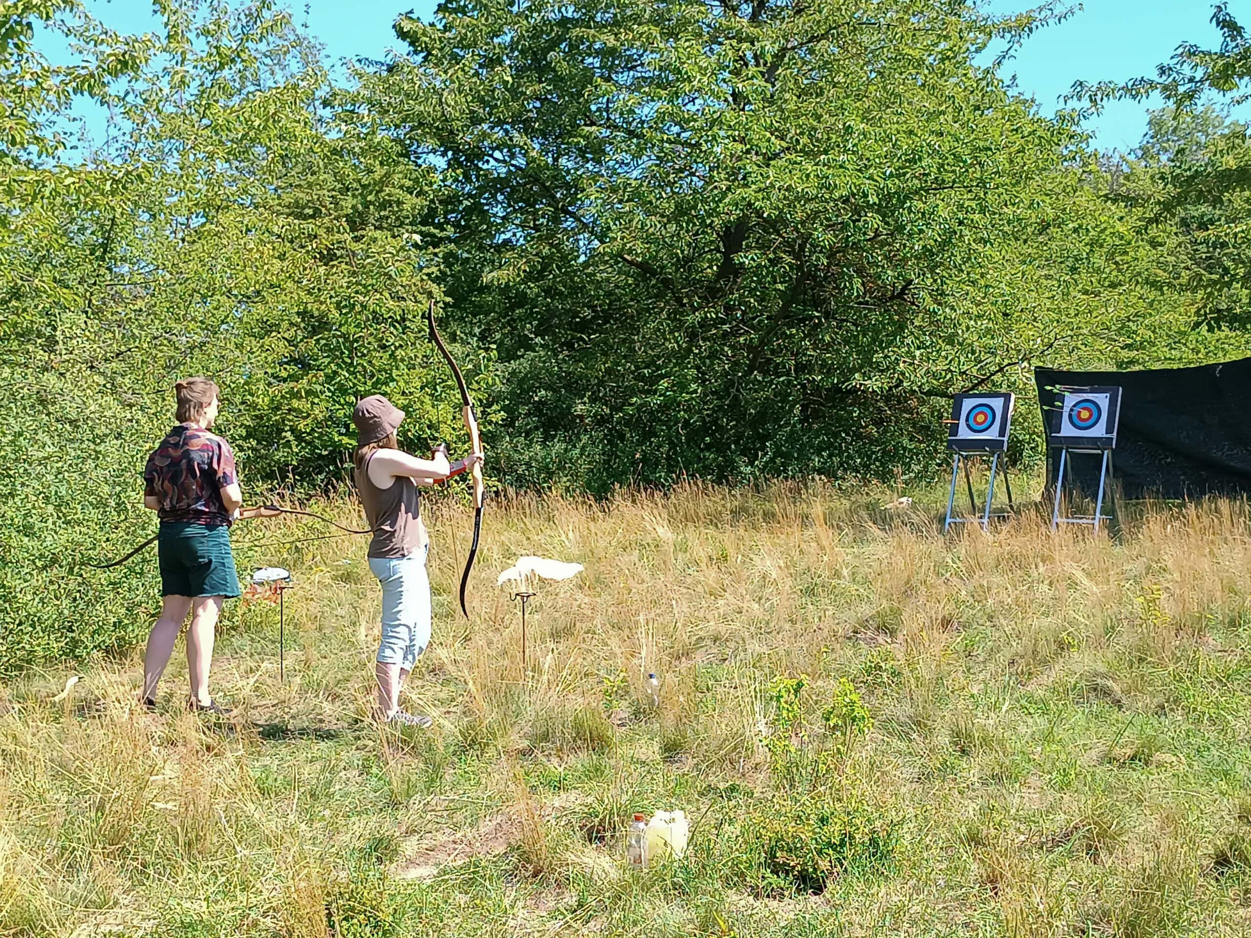 Susann und Justine beim Bogenschießen draußen auf eine Zielscheibe.