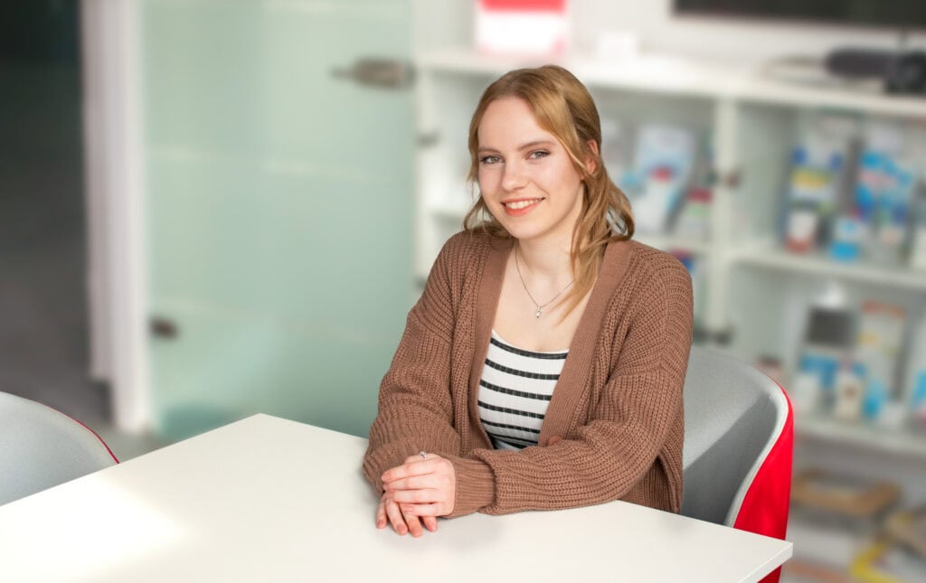 Isabel (Praktikantin) Unsere Praktikantin Isabel sitzt an einem Tisch bei uns im Meetingraum und lächelt in die Kamera.