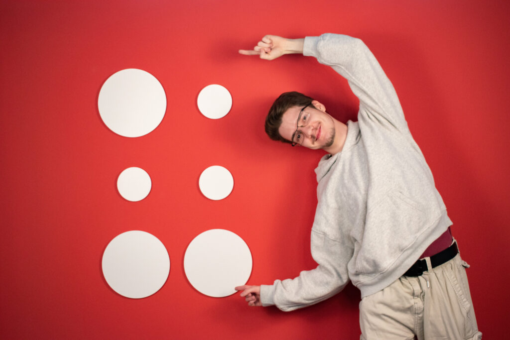 Praktikant Lukas steht vor roter Wand und zeigt auf das Hupa-Logo