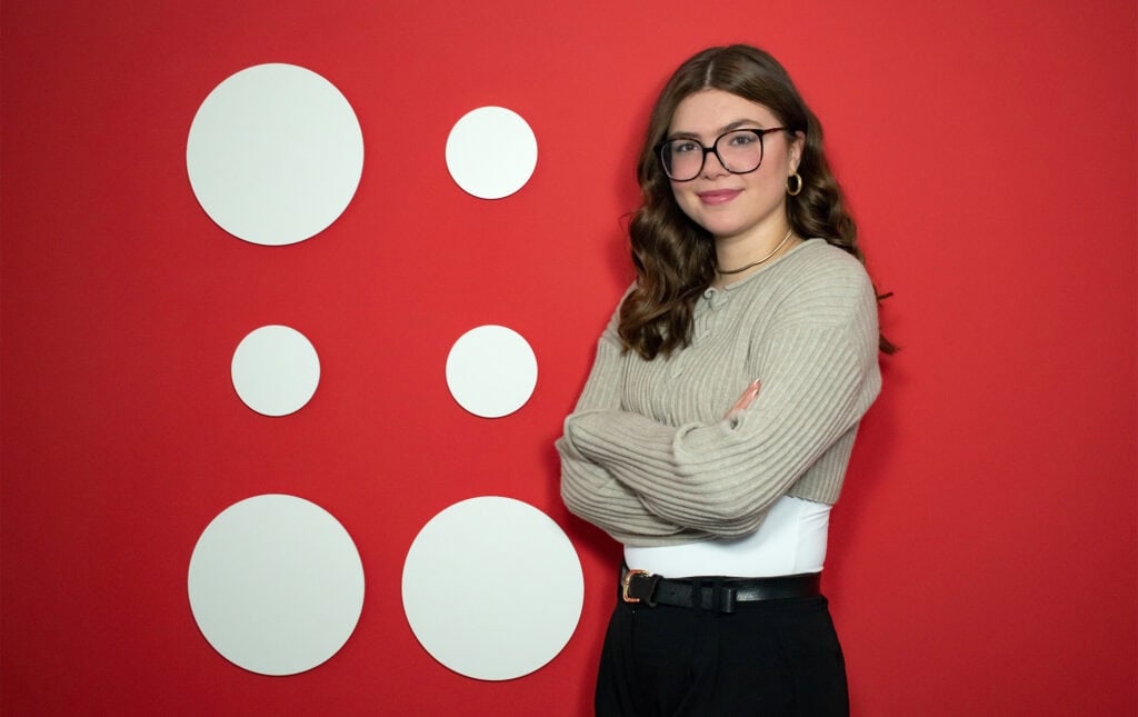 Praktikantin Michelle steht vor einer roten Wand mit dem Hupa Logo deauf