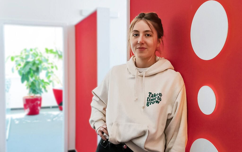 Praktikantin Luisa steht angelehnt an der roten Wand im Hupa Office neben dem Hupa Logo